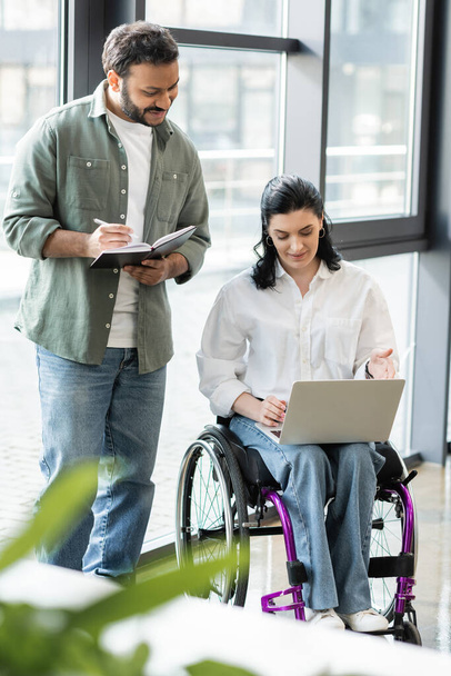 happy indian man taking notes near disabled businesswoman on wheelchair using laptop in office - Foto, Imagen