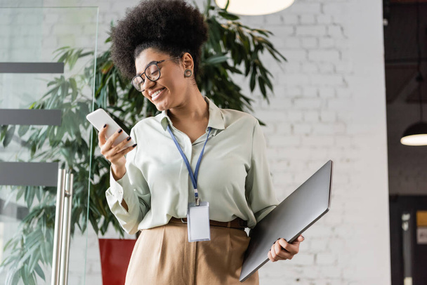 positive african american businesswoman in glasses holding laptop and using smartphone in office - Foto, Imagen