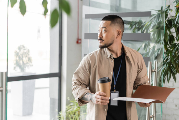 відволікаючий азіатський бізнесмен з значком, що йде в офіс з папкою і паперовою чашкою в руках - Фото, зображення