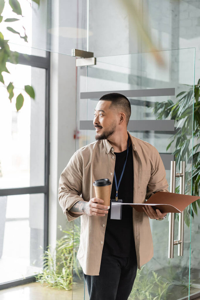 відволікаючий азіатський бізнесмен з бейджем посміхається і ходить в офіс з папкою і паперовою чашкою - Фото, зображення