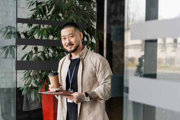 feliz asiático hombre de negocios con tatuaje sonriendo y de pie con carpeta y café en papel taza - Foto, Imagen