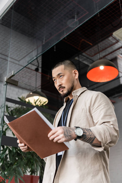 serious asian team leader with tattoo holding startup plan inside of folder near glass door - Photo, Image