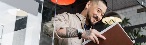 šťastný asijský podnikatel s tetováním při pohledu na dokumenty uvnitř složky u skleněných dveří, banner - Fotografie, Obrázek