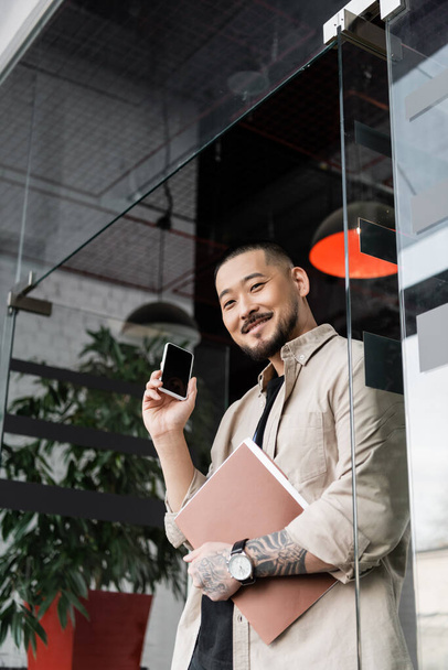 szczęśliwy azjatycki biznesmen z tatuaż gospodarstwa folder i smartfon w pobliżu szklane drzwi biura - Zdjęcie, obraz