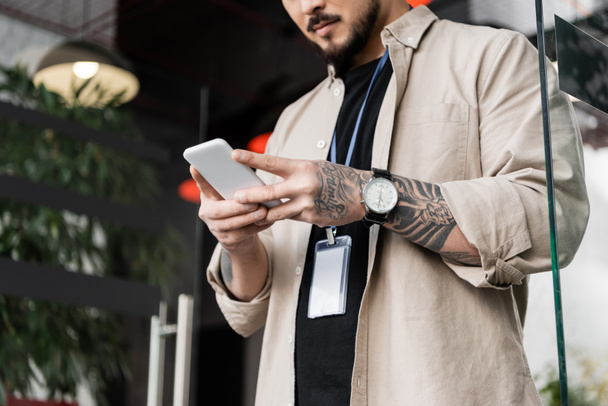 oříznutý pohled na vousatého podnikatele v neformálním oblečení, stojícího s odznakem a klábosícího na smartphonu - Fotografie, Obrázek
