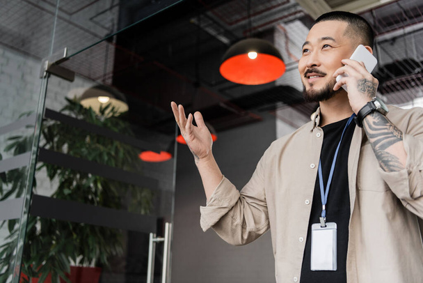 feliz asiático hombre de negocios con tatuaje gesto durante llamada de teléfono inteligente en la oficina moderna - Foto, imagen