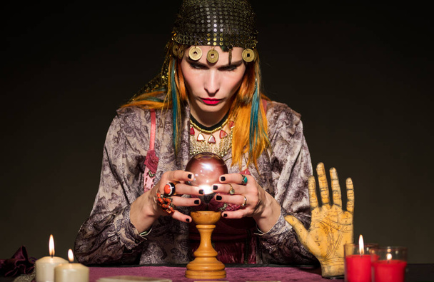 Εστιασμένη γυναίκα μάντης φορώντας αξεσουάρ κάθεται στο τραπέζι με αναμμένα κεριά και την τύχη λέει αντικείμενα, ενώ συμβουλεύονται κρυστάλλινη σφαίρα κατά τη διάρκεια της συνεδρίας μαντείας - Φωτογραφία, εικόνα