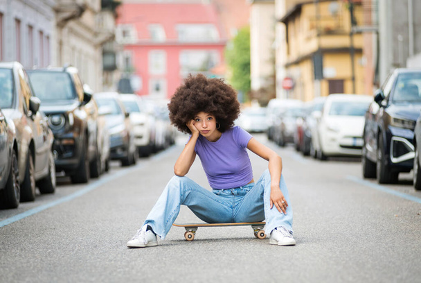 Przemyślana młoda Marokanka kobieta z afro włosy patrząc w aparat podczas siedzenia na deskorolce na ulicy w pobliżu zaparkowanych samochodów z ręką dotykając twarzy i opierając się na kolanie w świetle dnia - Zdjęcie, obraz