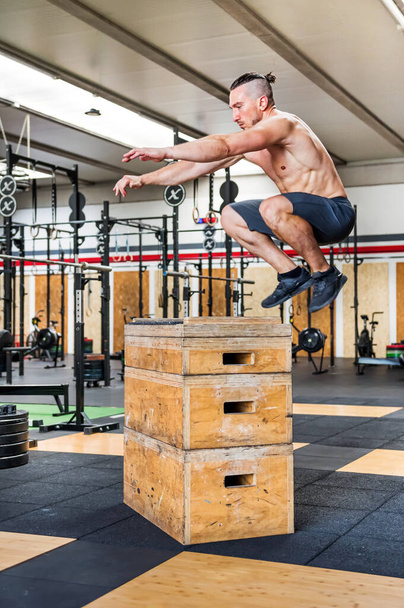 Vista lateral de cuerpo completo del atleta masculino muscular sin camisa haciendo ejercicio de salto de caja durante el entrenamiento intenso en el gimnasio - Foto, Imagen