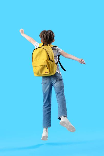 Стрибки маленького хлопчика зі шкільною сумкою на синьому фоні, вид ззаду - Фото, зображення