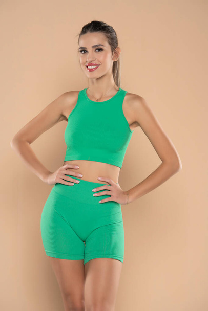 fiatal sportos mosolygós nő zöld rövidnadrágban és top pózol bézs stúdió háttér - Fotó, kép
