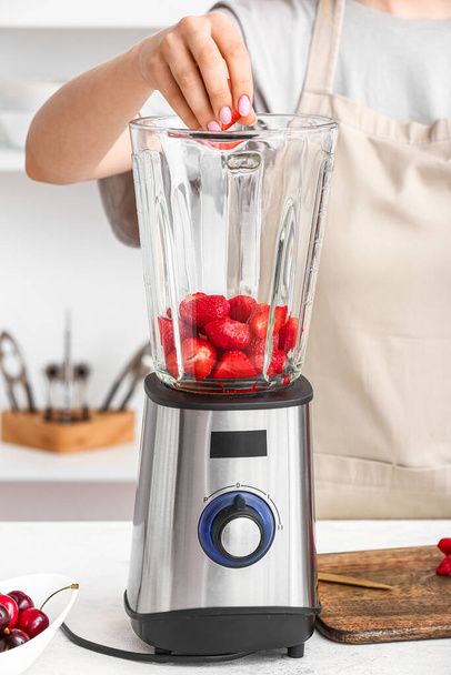 Woman preparing tasty strawberry smoothie with blender at table in kitchen - Photo, Image