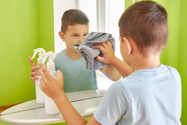 O menino lava o espelho com um pano e detergente. TPC. Limpeza da casa. - Foto, Imagem