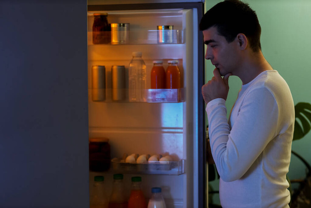 Nälkäinen nuori mies lähellä avointa jääkaappia keittiössä yöllä - Valokuva, kuva
