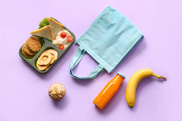 Bolsa, lonchera con deliciosa comida y botella de jugo sobre fondo lila - Foto, Imagen
