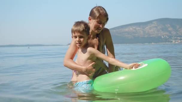 Joven madre cariñosa ayudando a su pequeño hijo a entrar en el anillo de natación inflable en el mar. Vacaciones familiares, vacaciones y divertido verano de niños y padres - Metraje, vídeo