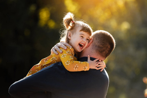 Papá alegre abraza a su pequeña hija sonriente. Papi soltero y el niño se divierten, ríen y disfrutan de la naturaleza al aire libre en el parque de otoño. Concepto de cuidado parental y feliz infancia sin preocupaciones. Feliz día de los padres. - Foto, Imagen