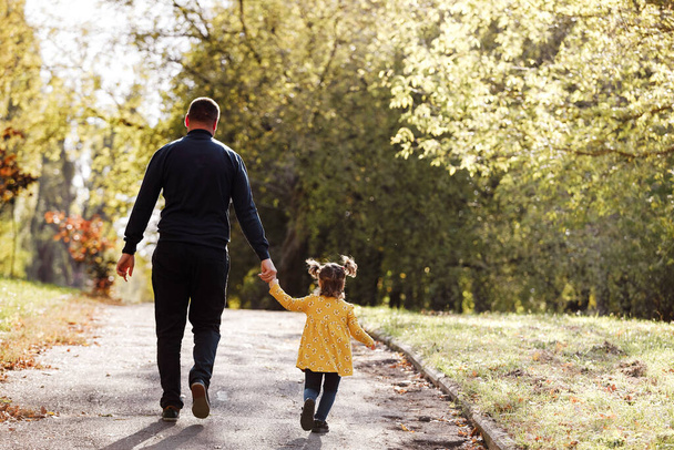 Happy fathers day. Rear view of dad holds hand his little daughter and walks outdoors at autumn park. Single daddy with child relaxes and spends time together. Family day and childhood concept - Photo, Image