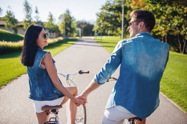 Back view of beautiful stylish couple talking and smiling while cycling in the park - Photo, Image