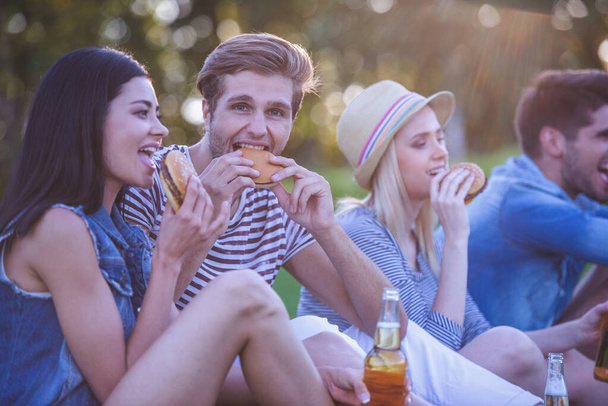 Hermosos amigos elegantes están bebiendo cócteles, comiendo sándwiches, comunicándose y sonriendo mientras descansan en el parque
 - Foto, imagen