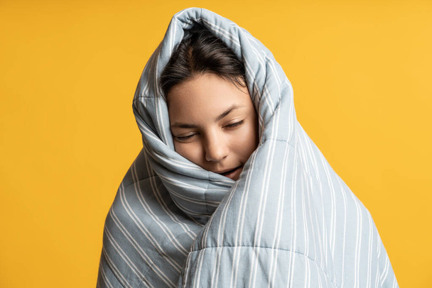 Ritratto di donna bruna assonnata avvolta in una coperta a righe grigie su sfondo giallo. Ragazza con gli occhi chiusi coperti di piumino caldo. Accogliente tempo di comfort, mancanza di sonno, pisolino, insonnia, disturbi del sonno. - Foto, immagini