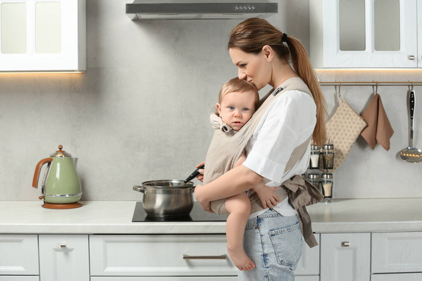 Mother holding her child in sling (baby carrier) while cooking in kitchen - Valokuva, kuva
