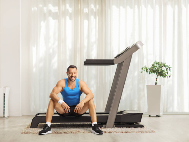 Smiling young man sitting on a treadmill at home and looking at camera  - Φωτογραφία, εικόνα