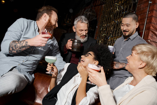 Африканская американка смеется рядом с многонациональными друзьями с напитками в баре, после рабочей вечеринки - Фото, изображение