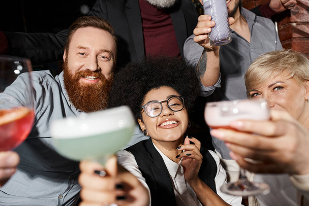 szczęśliwy brodaty mężczyzna i afrykański Amerykanin kobieta z koktajle patrząc w aparat w pobliżu kolegów w barze - Zdjęcie, obraz