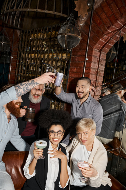 cheerful multicultural women looking at camera near bearded colleagues toasting in cocktail bar - Photo, Image