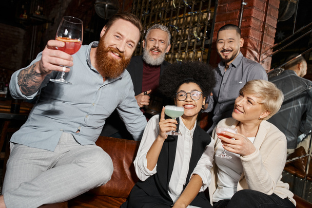 bärtiger tätowierter Mann mit Cocktail blickt in die Kamera in der Nähe multiethnischer Freunde, die sich in einer Bar amüsieren - Foto, Bild