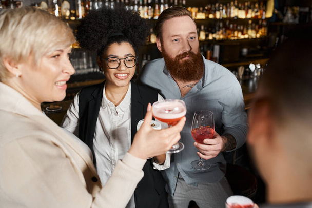 bearded tattooed man and african american woman smiling near workmates with cocktail glasses in bar - Foto, Imagem