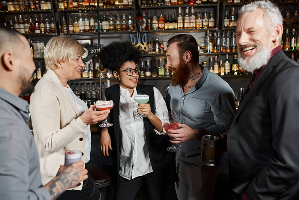 Sakallı, dövmeli adam, barda içki içip iş arkadaşlarının yanında gülümseyen Afrikalı Amerikalı bir kadınla konuşuyor. - Fotoğraf, Görsel