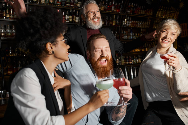 hombres barbudos felices riendo cerca de mujeres multiculturales en el bar de cócteles, diversa fiesta de equipo después del trabajo - Foto, Imagen