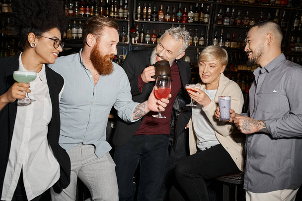татуированный бородатый мужчина держит стекло возле веселых мультиэтнических коллег в баре, после досуга - Фото, изображение