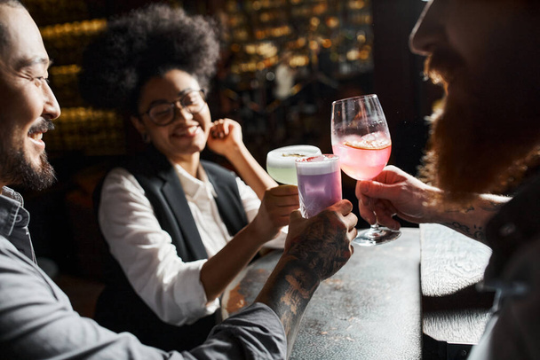 sorridente donna afro-americana con occhiali da cocktail e colleghi tatuati barbuti nel bar - Foto, immagini