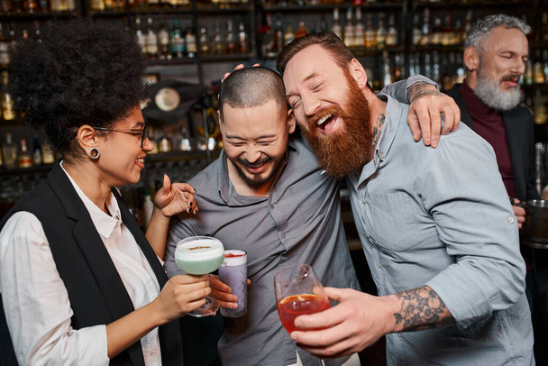 tattooed multiethnic colleagues with cocktails laughing near cheerful african american woman in bar - Photo, Image