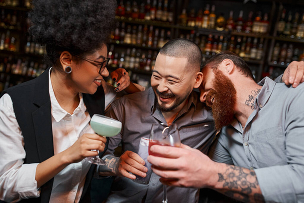 überglückliche multiethnische Arbeitskollegen mit Cocktails, die sich umarmen und lachen in der Bar, Freizeit und Spaß - Foto, Bild