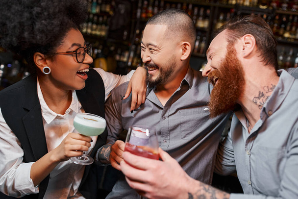 excited multiethnic colleagues with drinks embracing and laughing in bar, leisure after work - Photo, Image