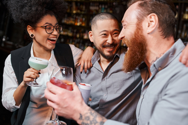 fröhliche multiethnische Arbeitsfreunde mit Gläsern, die sich nach Feierabend in der Cocktailbar umarmen und lachen - Foto, Bild