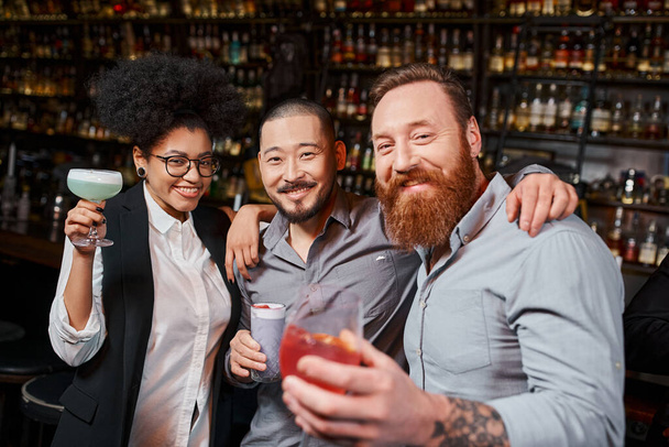 colegas multiétnicos segurando óculos com coquetéis e sorrindo para a câmera no bar, descansar depois do trabalho - Foto, Imagem