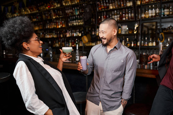 vrolijk aziatische man en afrikaanse amerikaanse vrouw met drankjes glimlachen naar elkaar in cocktailbar - Foto, afbeelding