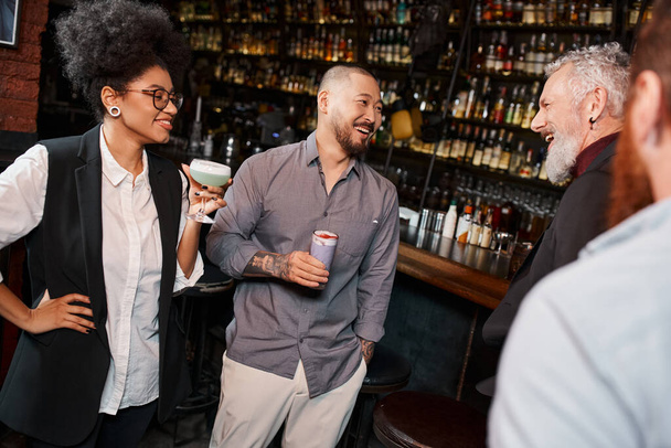 Afrykańska amerykanka z koktajlem uśmiechnięta obok brodatych, wielonarodowych kolegów rozmawiających w barze - Zdjęcie, obraz