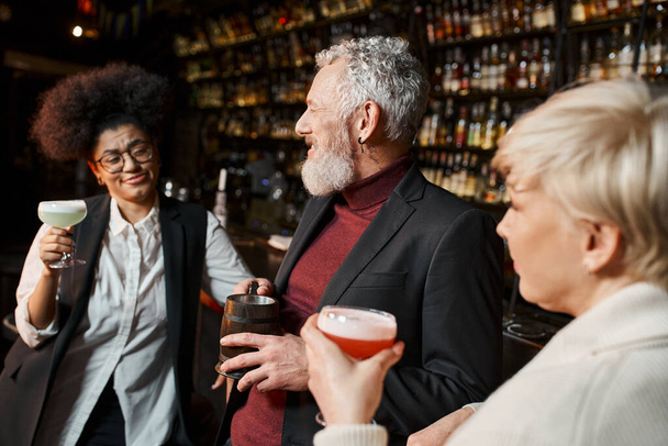 бородатый мужчина средних лет улыбается рядом с многонациональными женщинами с коктейлями, коллеги отдыхают в баре - Фото, изображение