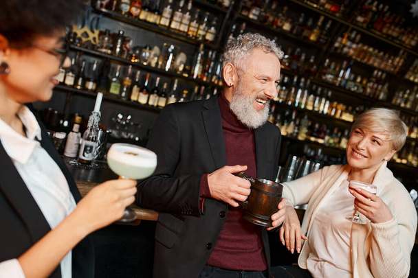 multiethnic women holding glasses and looking at cheerful bearded colleague in cocktail bar - Photo, Image