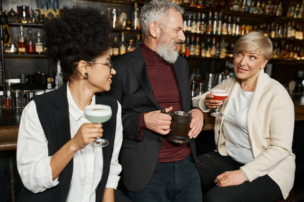 wielonarodowe kobiety w okularach koktajlowych uśmiechnięte podczas rozmowy z brodatym kolegą w barze - Zdjęcie, obraz