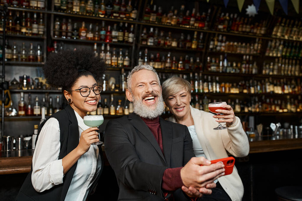 alegre hombre barbudo tomando fotos con mujeres multiétnicas en el bar, diverso equipo descansando después del trabajo - Foto, Imagen