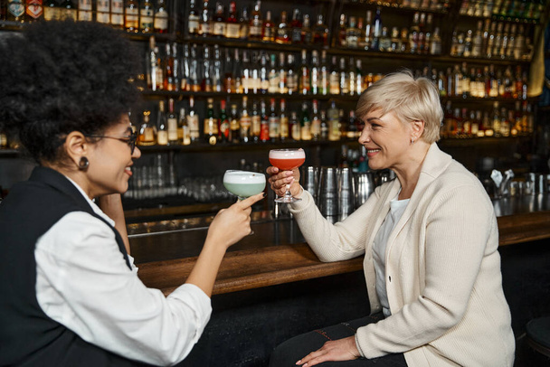 smiling multiethnic women toasting with cocktails glasses while spending time in bar after work - Photo, Image