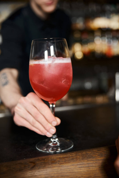 καλλιεργημένη άποψη του μπάρμαν κρατώντας γυαλί με κοκτέιλ φρούτων αλκοόλ στο μπαρ, επαγγελματική mixologist - Φωτογραφία, εικόνα