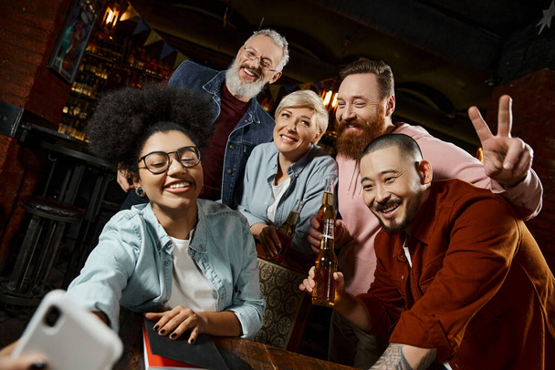 cheerful african american woman taking photo with multiethnic colleagues holding beer bottles in pub - Photo, Image